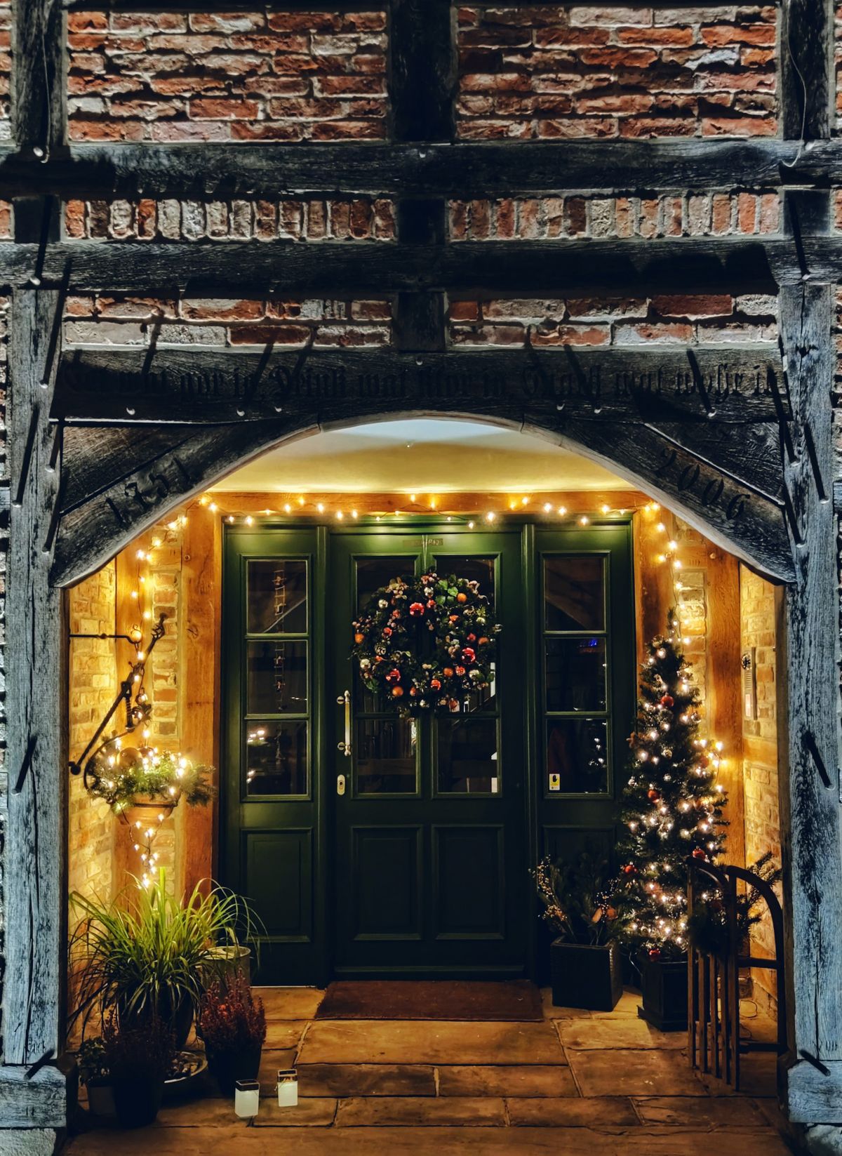 Front door with a lit wreath, garland around the arch and a small lit tree beside it